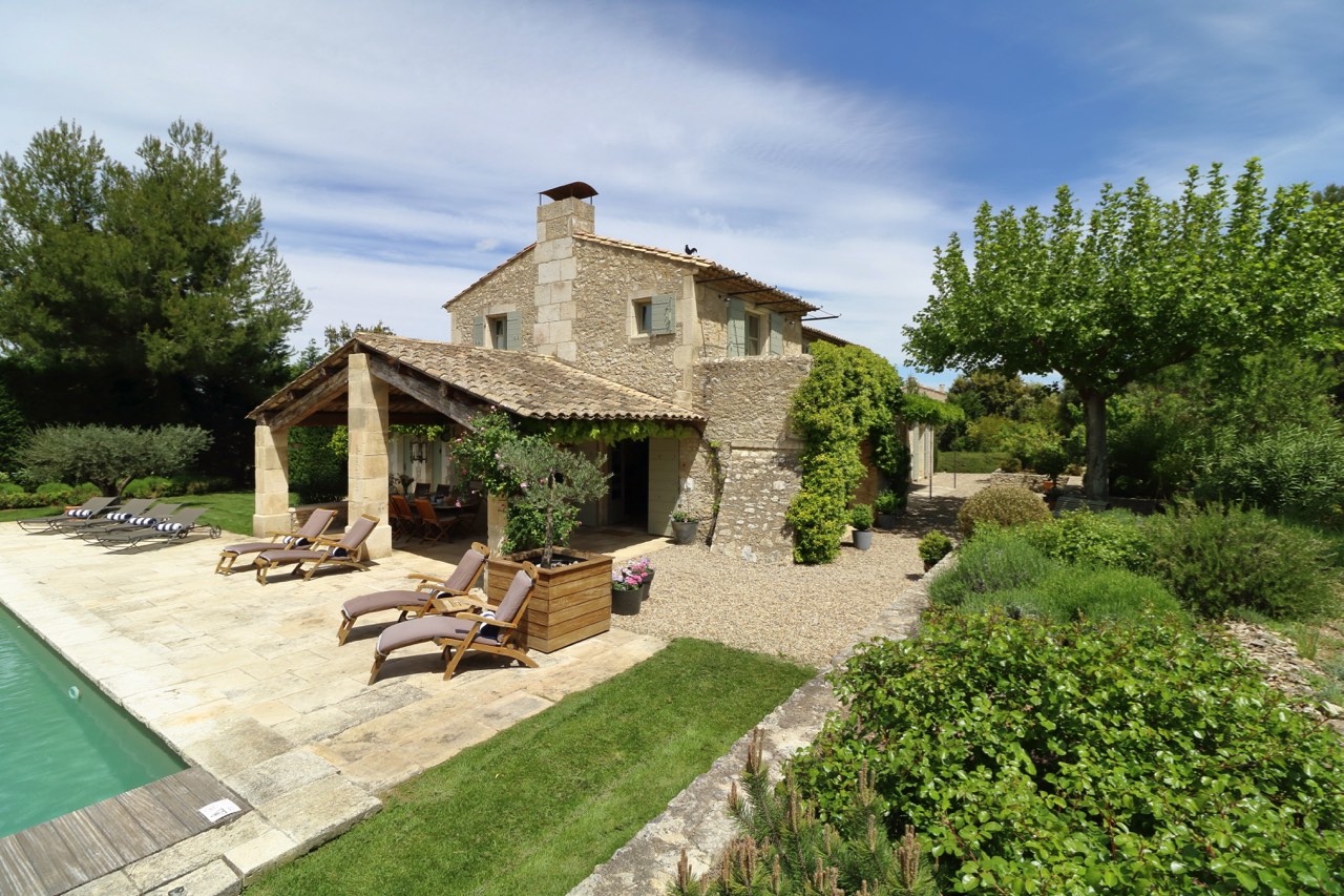 A charming stone villa with a covered patio, next to a swimming pool and surrounded by lush greenery under a clear blue sky.